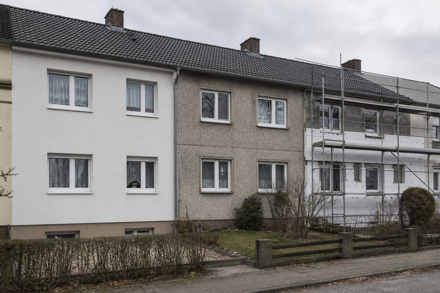 Drei aneinandergebaute Reihenhäuser in unterschiedlichem Sanierungszustand: links neu gedämmt und verputzt, mittig alter Kratzputz mit alten Fenstern, rechts eingerüstet mit teilweise montierter Dämmung