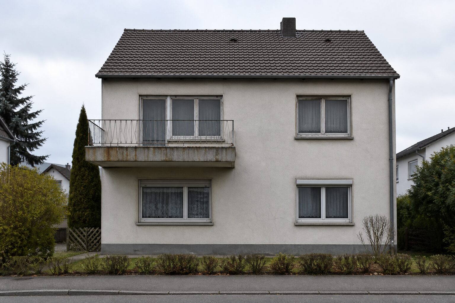 Straßenansicht eines Einfamilienhauses der 1960er Jahre mit Putzrissen, alten Fenstern, rostfleckiger Balkonplatte und verschobenen Dachpfannen