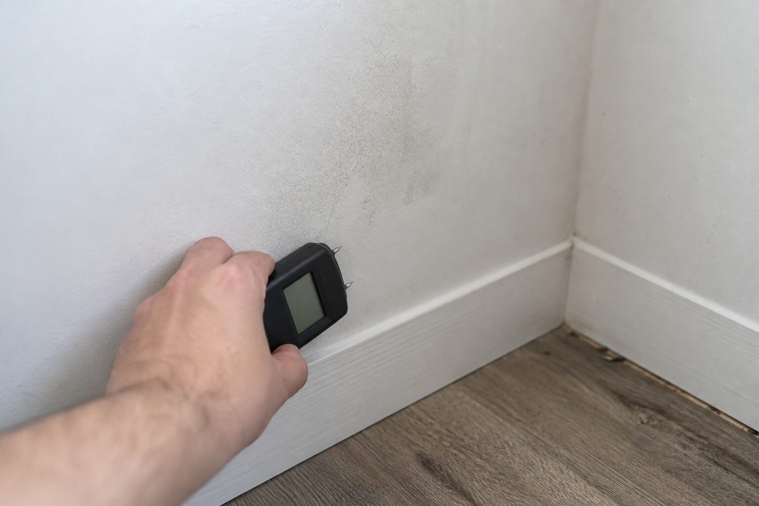 Feuchtemessgerät wird an einen dunklen Feuchtefleck über der Sockelleiste gehalten