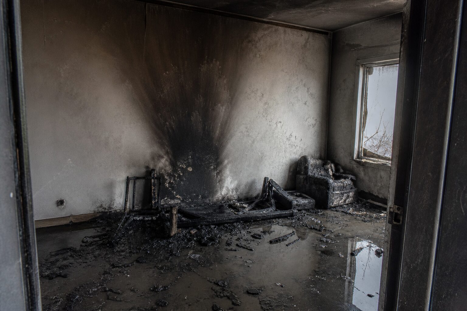 Ausgebrannter Wohnraum mit Rußfahne an der Wand, verkohlten Möbelresten und Löschwasser auf dem Boden