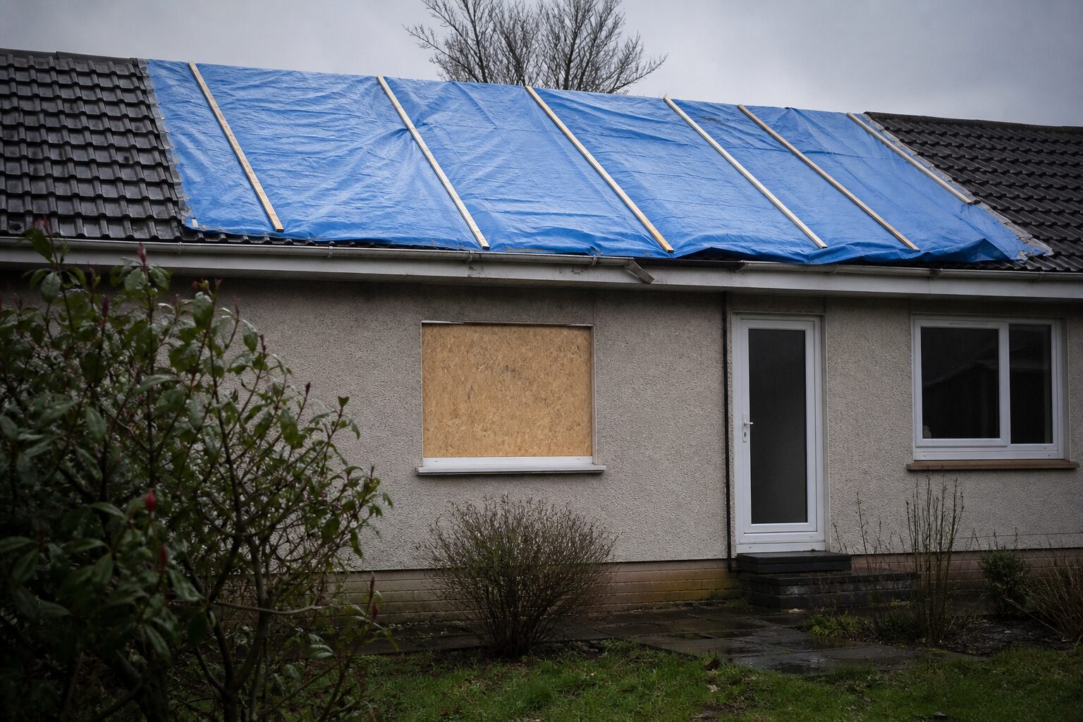 Wohnhaus mit blauer Notabdeckung über der beschädigten Dachfläche und einem mit Platte verschlossenen Fenster